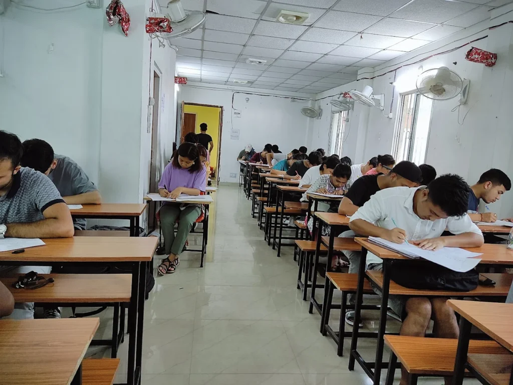 The image shows an indoor classroom setting where numerous students are seated at individual desks, diligently writing on papers. The room is brightly lit with fluorescent lights, and several ceiling fans are visible. The walls are painted a light color, and a doorway is visible at the far end of the room.