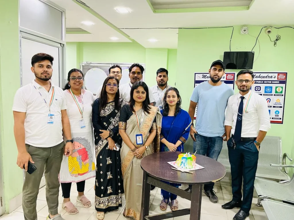 A group of ten individuals is captured in a brightly lit indoor setting, likely an office, as they pose for a photograph around a small, round table adorned with a colorful birthday cake. The group comprises six women and four men, all seemingly in good spirits.
The woman positioned centrally in the front row stands out; she is clad in a light brown or beige saree draped over a white blouse and secured with a safety pin at the shoulder. She wears a lanyard around her neck holding an identity card. To her left, another woman is dressed in a white kurta featuring a vibrant, abstract design in red, yellow, and blue hues, paired with dark leggings. The woman to the right of the saree-clad individual is wearing a royal blue kurta with matching pants. The remaining women in the back row are dressed in a mix of traditional and Western attire, including a black and white patterned kurta.
The men are dressed in casual and semi-formal wear. One man on the left sports a white t-shirt with a green, white, and orange ribbon pinned to it, along with khaki pants. Another man in the back row wears a light blue collared shirt. To the right, a man is seen in a light blue t-shirt and jeans, while the man on the far right is dressed in a crisp white shirt, a dark tie with white stripes, and dark trousers. Most individuals in the photograph are wearing lanyards with identification cards.
The backdrop of the image features light green walls, a glass-paneled door, and a whiteboard. On the right side, a poster or signage with text and logos, including what appears to be "Mahendra's," is visible. The overall ambiance suggests a celebratory occasion, likely a birthday, within a professional environment.