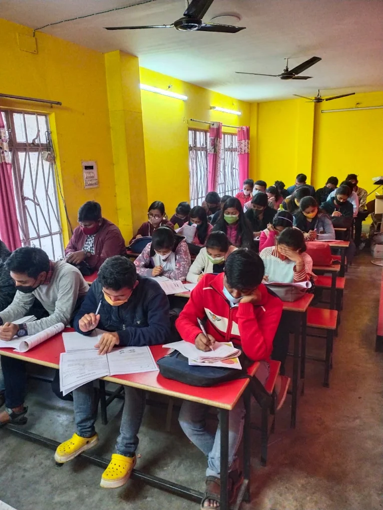 The image shows a brightly lit classroom in Siliguri, West Bengal, India, filled with students diligently working at their desks. The walls of the classroom are painted a vibrant yellow. Most of the students are wearing face masks. Each student is seated at an individual desk with a red tabletop and a metal frame, working on papers. Ceiling fans are visible above. Natural light enters the room through a window on the left side, which has metal bars and red curtains. The overall atmosphere is focused and studious.