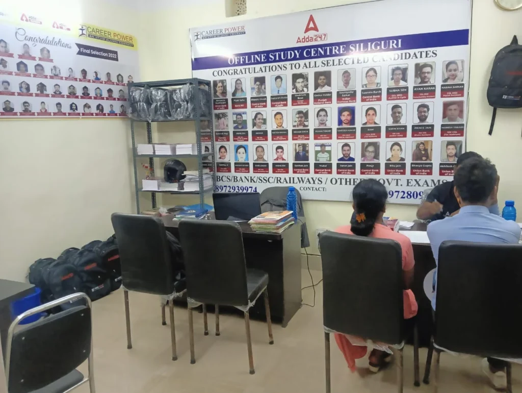 The image shows an indoor setting with a group of people posing in front of a banner. The banner features the logos "CAREER POWER" and "Adda 24.7," and includes photos of individuals with text congratulating selected candidates. A desk with books and some chairs are visible in the foreground.