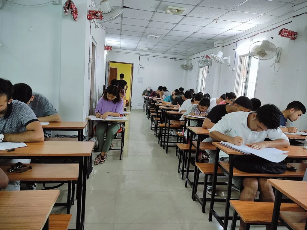 The image shows an indoor classroom setting where numerous students are seated at individual desks, diligently writing on papers. The room is brightly lit with fluorescent lights, and several ceiling fans are visible. The walls are painted a light color, and a doorway is visible at the far end of the room.