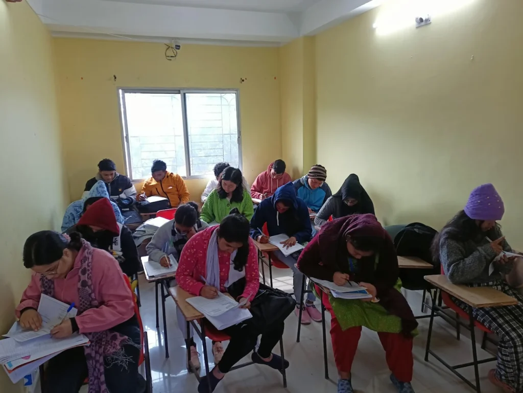 The image depicts an indoor classroom setting with numerous students seated at individual desks, focused on writing. The room is brightly lit by fluorescent lights, with several ceiling fans visible. The walls are painted a light color, and a doorway is visible at the far end of the room.