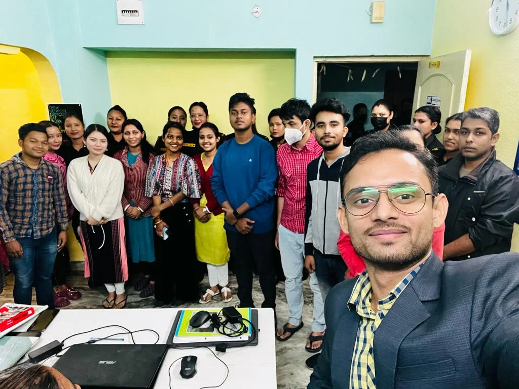 The image shows a group of about fifteen people posing for a selfie in what appears to be an office or community space. The group includes both men and women, who seem to be of South Asian descent, given the clothing and the location context. They are dressed in a mix of casual and traditional Indian attire. The person taking the selfie is a man in a suit jacket and glasses. The room has light green walls and a doorway is visible in the background. A laptop is placed on a table in the foreground.