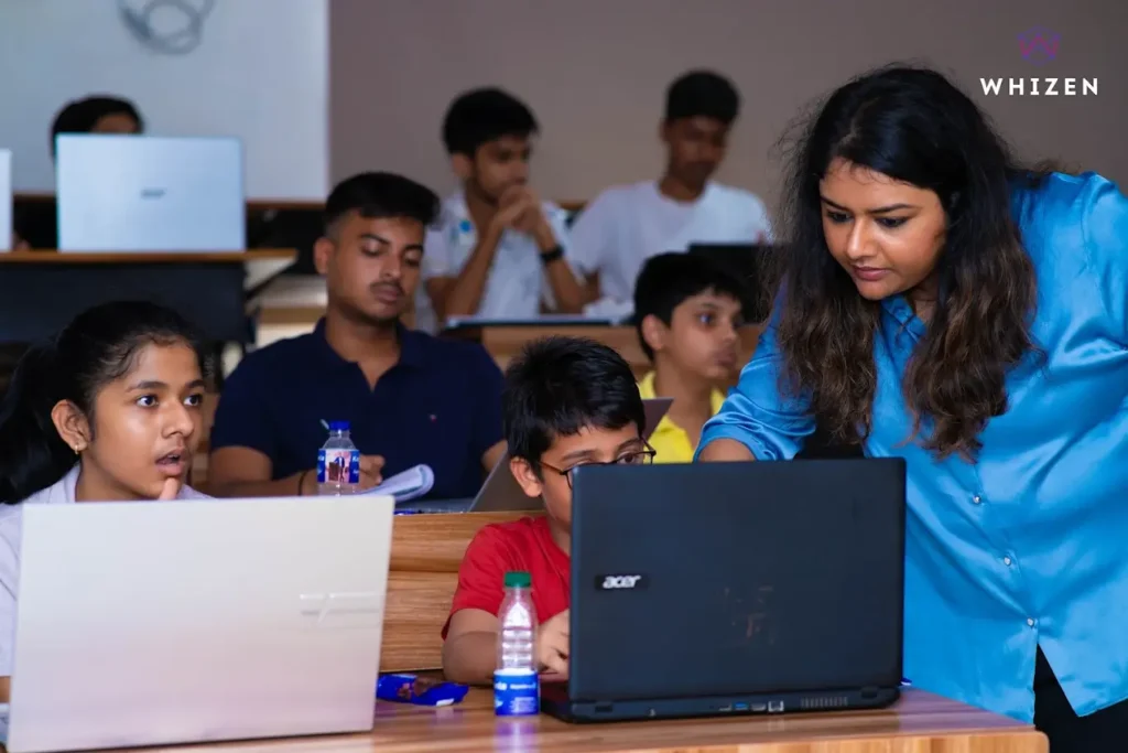 students sitting with laptops while a teacher guides them through a lesson