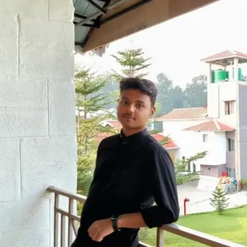 A young man with short dark hair, wearing a black long-sleeve shirt and light-colored pants, leaning casually against a metal balcony railing. He is standing next to a white brick column, and the background features a green lawn, trees, and buildings with red-tiled roofs.
