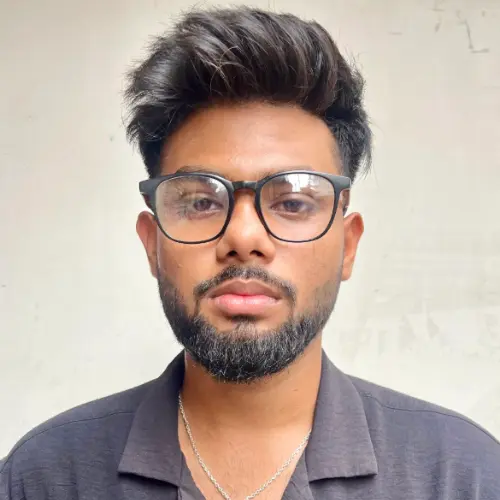 A close-up portrait of a young man with a beard and styled hair, wearing black-rimmed glasses, a black collared shirt, and a silver chain necklace.