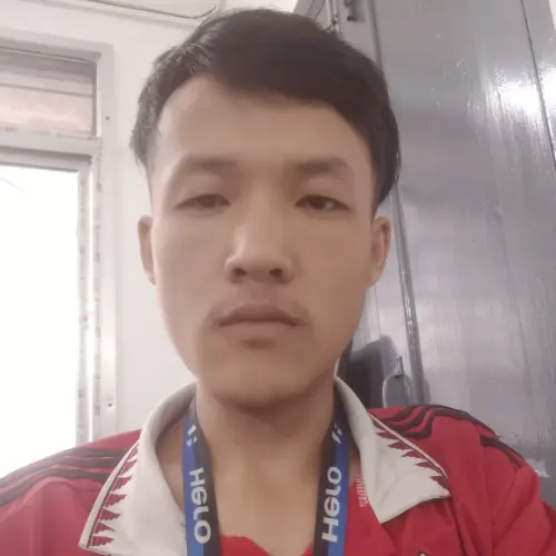 A close-up portrait of a young man with a beard and styled hair, wearing black-rimmed glasses, a black collared shirt, and a silver chain necklace.