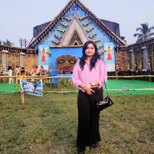 A full-body shot of a young woman with long dark hair wearing a pink shirt and black pants, standing on a grassy field in front of a large, elaborate blue and bamboo festival structure.