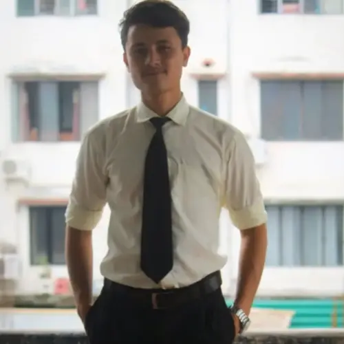 A young man wearing a white dress shirt with a black tie and black pants, standing with his hands in his pockets in front of a white building with windows.