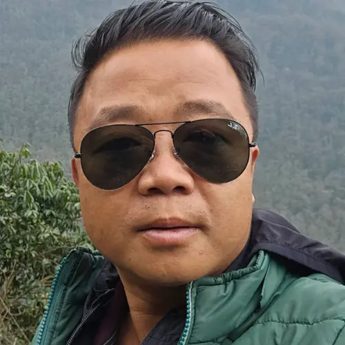 A close-up outdoor selfie of a man with short dark hair wearing aviator sunglasses and a green jacket, with a blurred background of trees and hills.