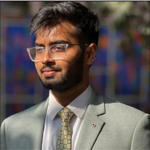 A sunlit outdoor portrait of a young man with a beard and clear rectangular glasses, wearing a light grey suit and a patterned tie, looking to the side against a blurred colorful background.