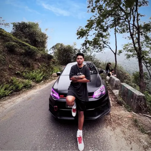 A full-body shot of a young man wearing a black t-shirt and white sneakers, leaning with his arms crossed against a black car with pink headlights on a hillside road.