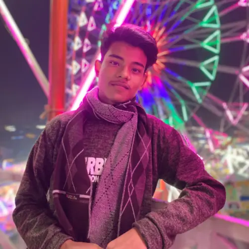 A young man wearing a grey argyle scarf and a dark jacket standing at night in front of a colorful, illuminated Ferris wheel.