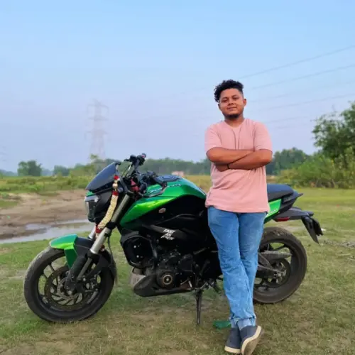 A young man with curly dark hair, wearing a pink t-shirt and blue jeans, standing with his arms crossed next to a green and black motorcycle in a grassy field.