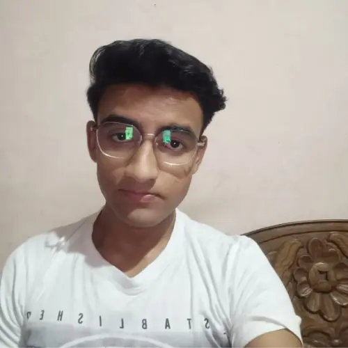A close-up indoor selfie of a young man with short dark hair and wire-rimmed glasses, wearing a white t-shirt with reversed text, sitting in front of a carved wooden chair back.