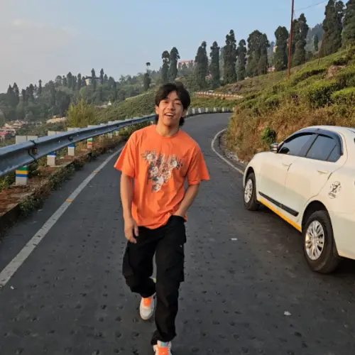 A full-body shot of a smiling young man walking on a paved road lined with tea gardens, wearing an orange graphic t-shirt and black cargo pants, with a white car parked on the side.