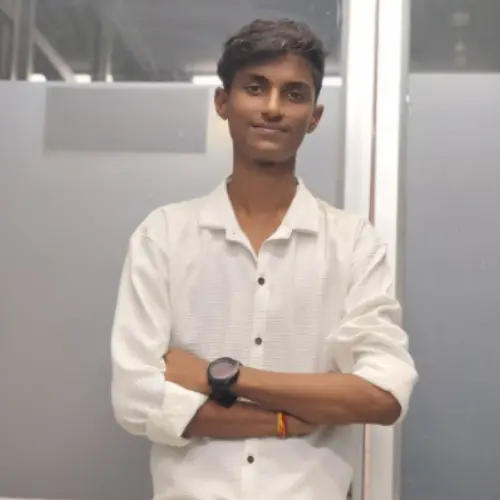 A portrait of a young man with short dark hair, wearing a white shirt and a black wristwatch, standing with arms crossed in front of a frosted glass partition.