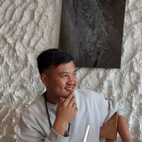 A portrait of a young man with short dark hair and a light mustache, wearing a grey sweatshirt and a beaded necklace, sitting against a textured white wall while smiling and looking to the side.