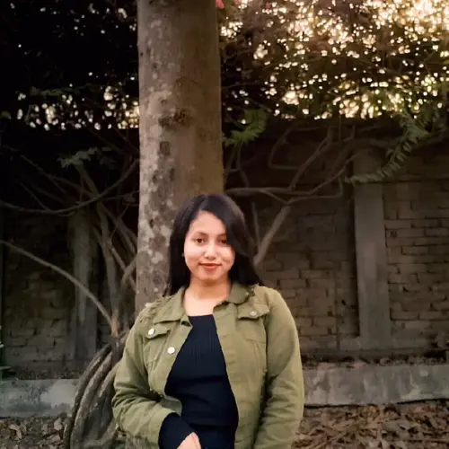 A medium shot of a young woman with long dark hair wearing a green jacket over a black top, standing outdoors next to a tree trunk with a brick wall in the background.