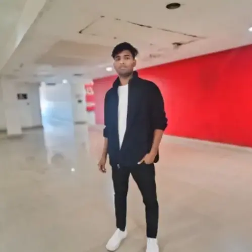 A full-body indoor shot of a young man wearing a dark jacket over a white t-shirt and black pants, standing in a hallway with a bright red wall on the right side.