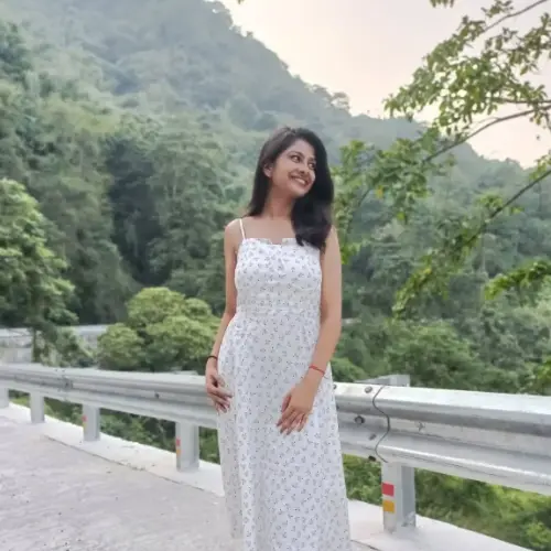 A full-body outdoor shot of a smiling young woman wearing a white patterned sundress, standing on a bridge with a metal railing against a backdrop of lush green hills.