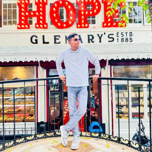 A full-body outdoor shot of a young man wearing sunglasses and a light grey hoodie, leaning against a black railing in front of a white building with a large red "HOPE" sign and "GLENARY'S" text.