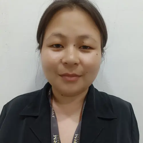 A close-up portrait of a young woman with dark hair pulled back, wearing a black blazer and a lanyard, standing against a plain white wall.