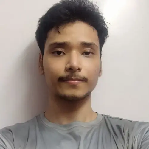 A close-up portrait of a young man with short dark hair and a mustache, wearing a grey t-shirt and looking directly at the camera against a plain white wall.