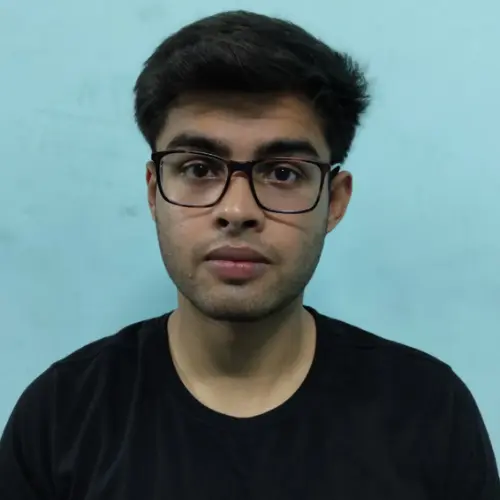 A close-up portrait of a young man with short dark hair and black rectangular glasses, wearing a black t-shirt and looking directly at the camera against a plain light blue background.