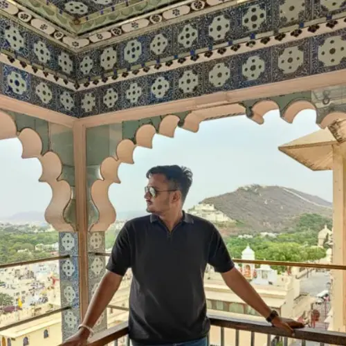 a man standing on a balcony or terrace with decorative, traditional archways. He is wearing a dark polo shirt and sunglasses, looking out toward a view that includes a hill and cityscape in the background.
