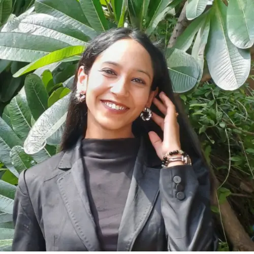 A smiling woman with long dark hair wearing a black blazer and silver hoop earrings, touching her hair while standing in front of large green leaves.