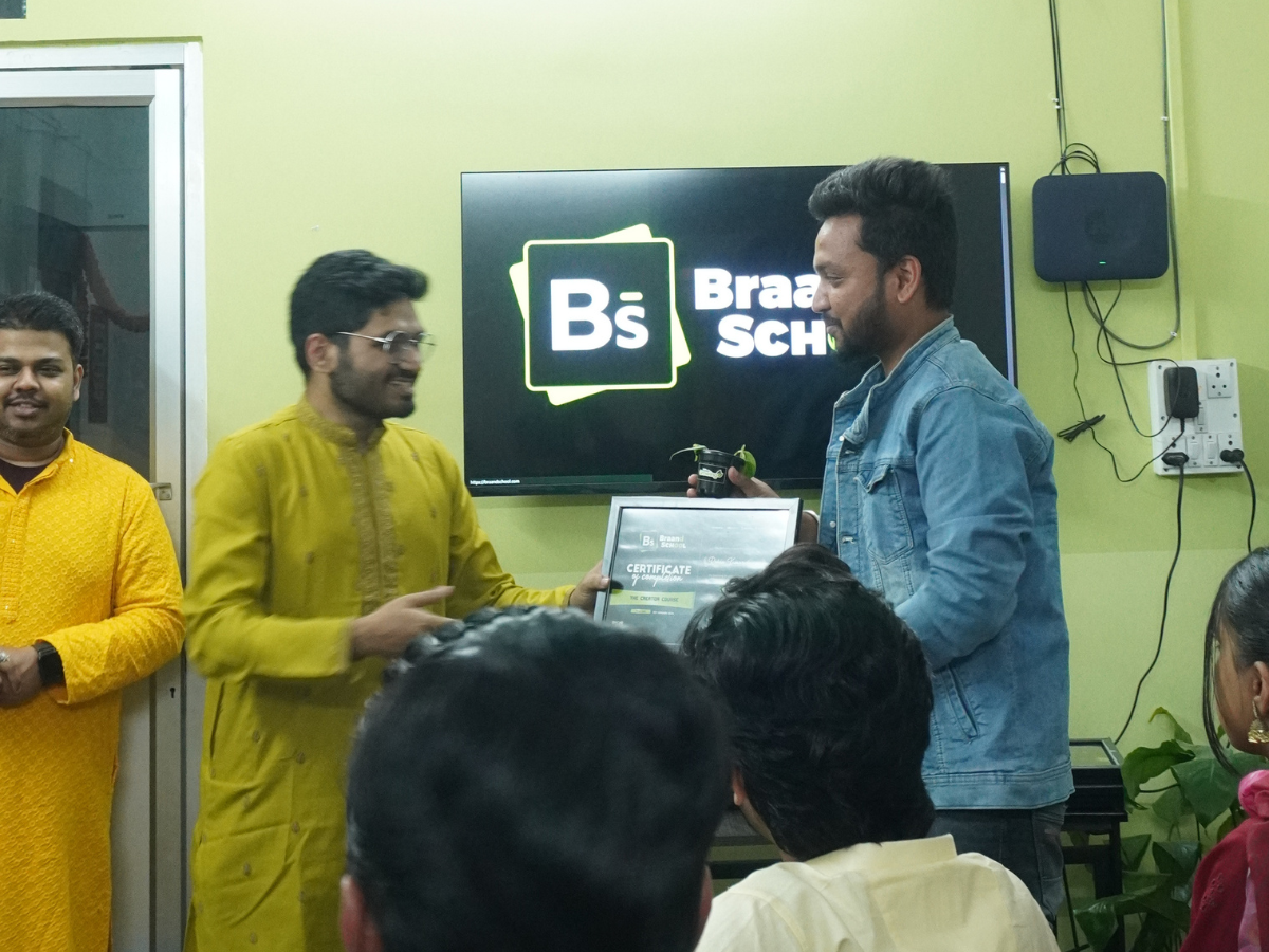 A student in a denim jacket receiving "The Creator Course" certificate and a plant gift from a mentor in a yellow kurta.
